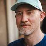 Close-up portrait of a man wearing a light blue cap and blue shirt, captured outdoors.